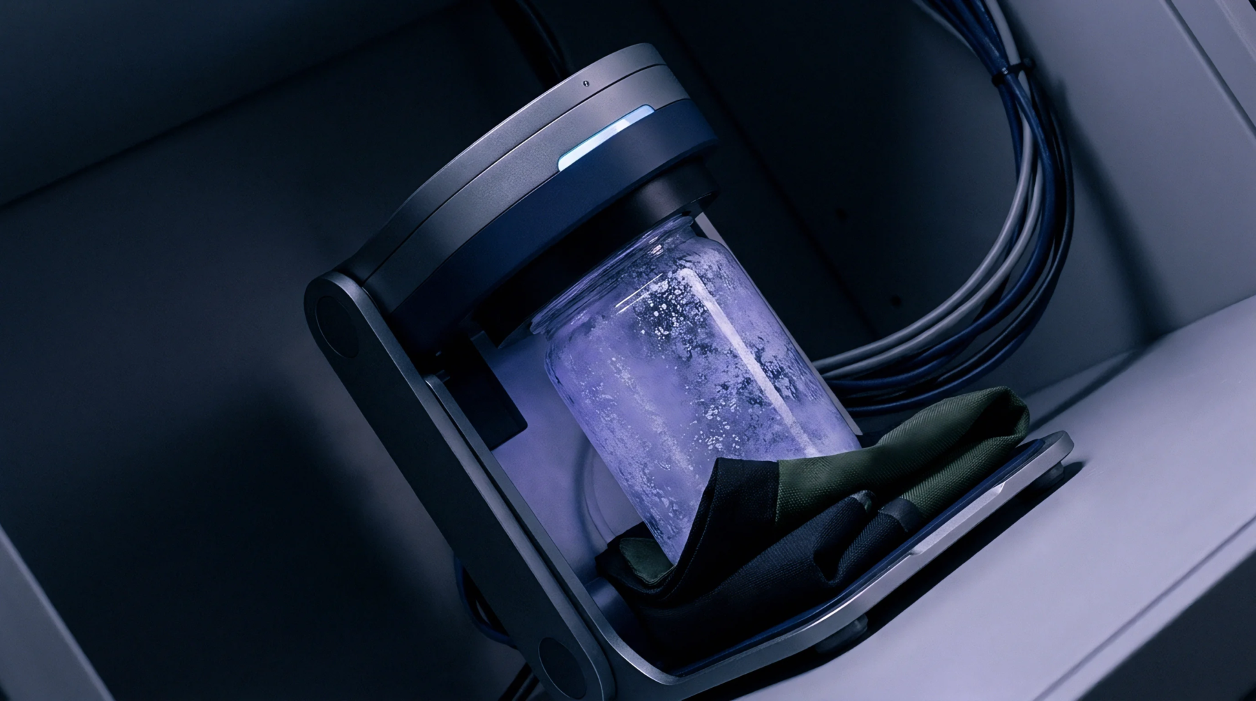 An automated robotic arm assembly holds a translucent glass specimen container above a dark fabric cloth within an enclosed technical instrument chassis.